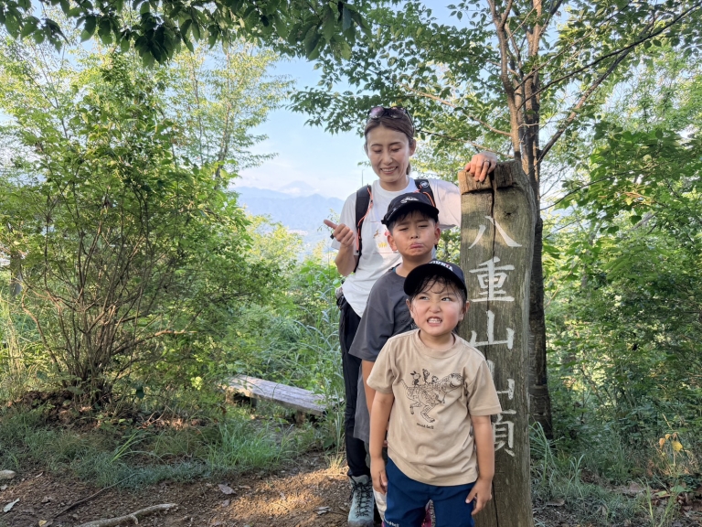 八重山山頂の標識前で上野原の山並みを背にする親子