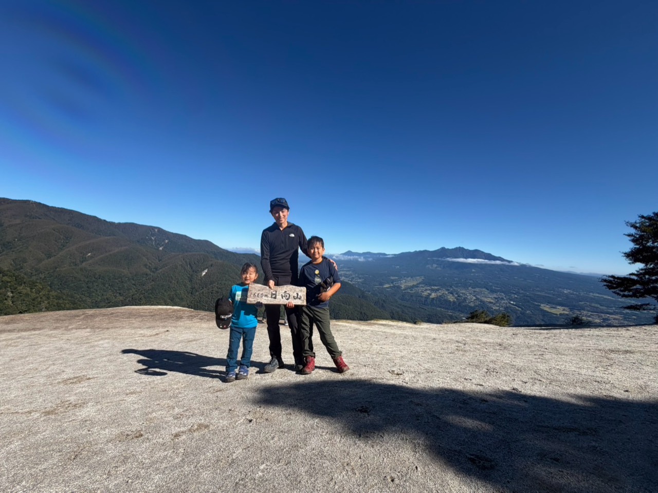 日向山の天空のビーチで家族登山 日向山の白砂と山並みを背景に山頂標識を持つ親子3人