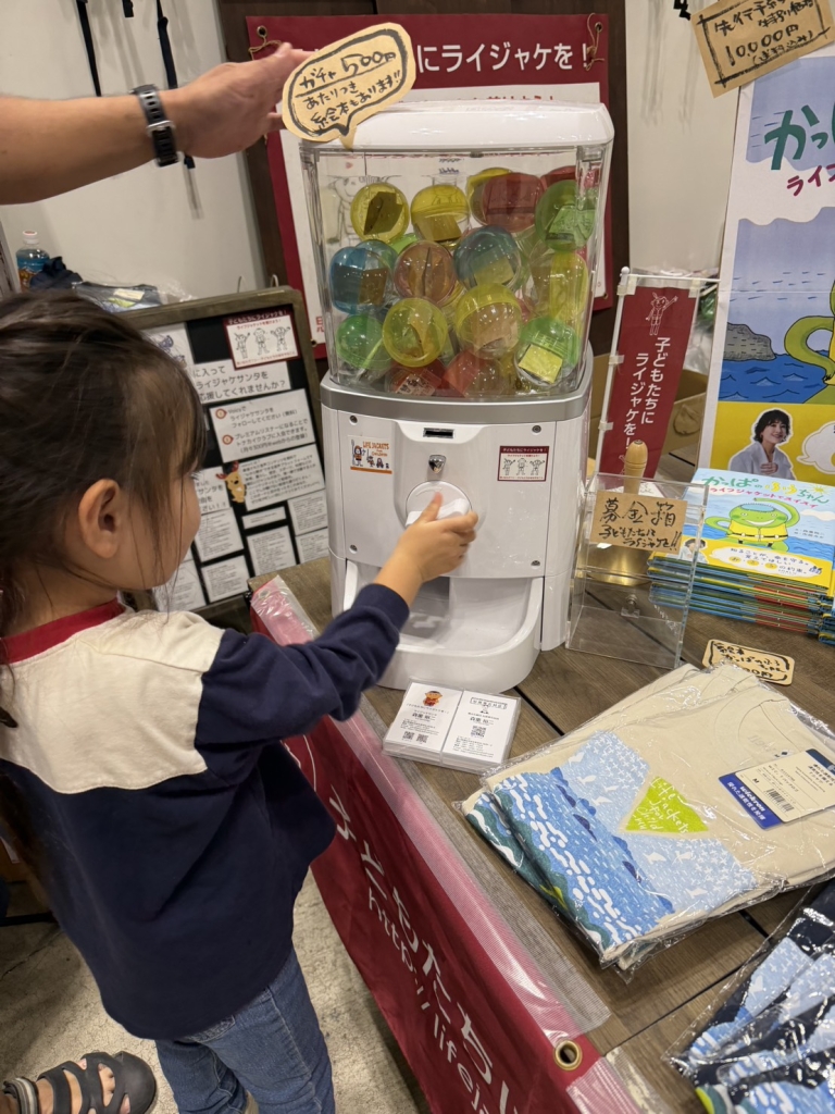 子どもたちにライジャケをのブースでカプセルトイを回す娘
